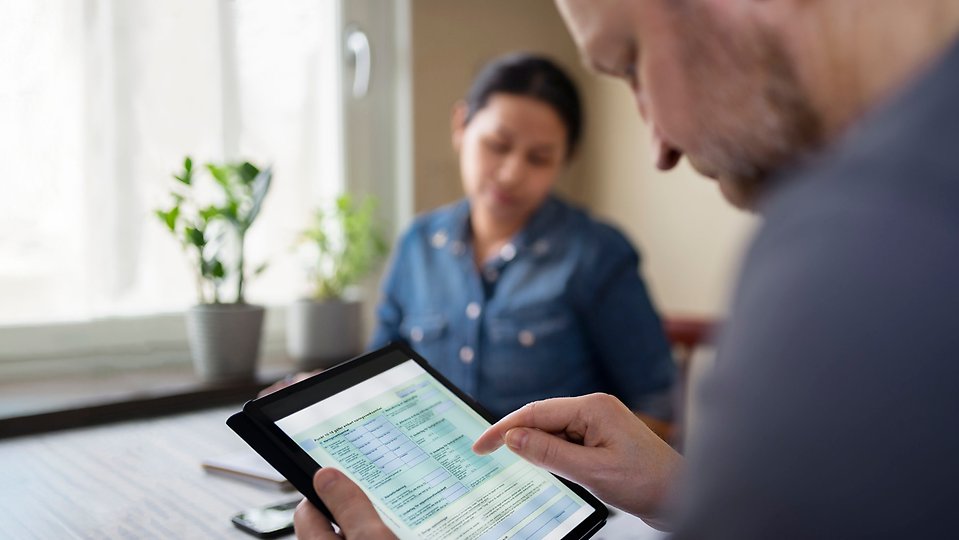 Person visar ett formulär på en surfplatta, med en annan person i bakgrunden.